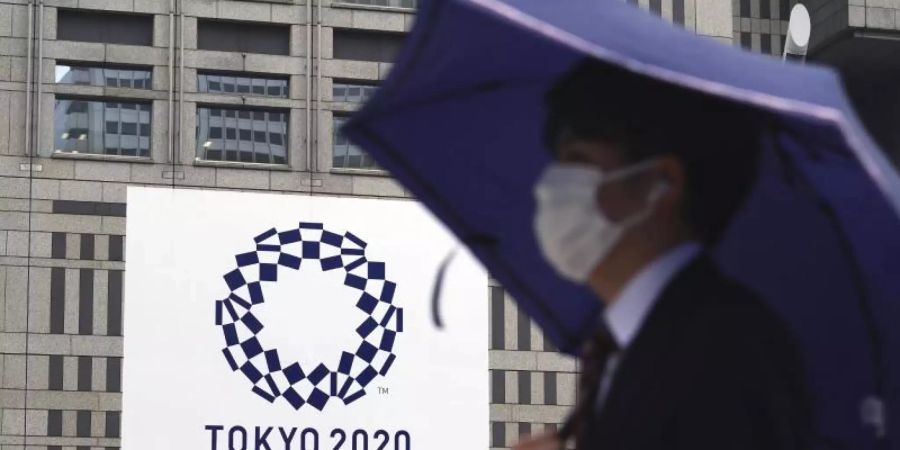 Ein Mann mit Mund-Nasen-Schutz geht im Regen an einem Banner der Olympischen Spiele Tokio 2020 vorbei. Foto: Eugene Hoshiko/AP/dpa