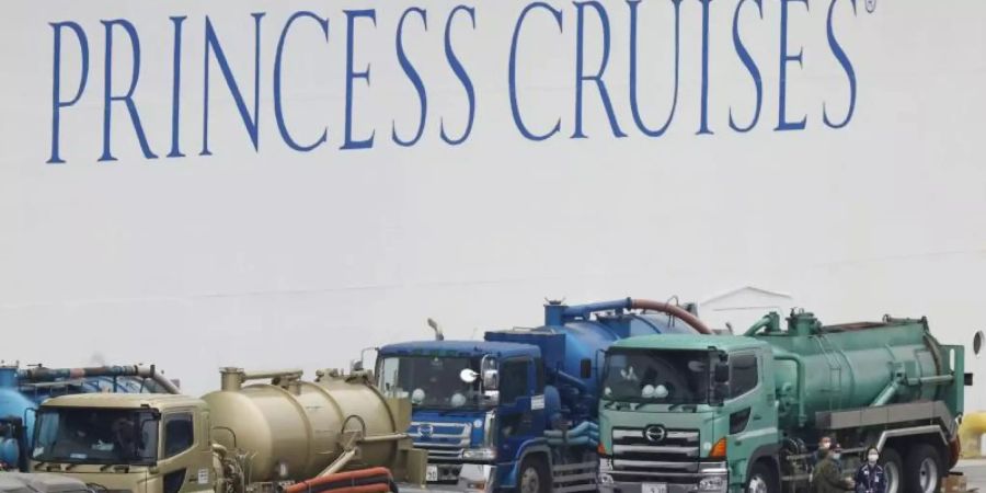 Menschen mit Mundschutz und teilweise mit Schutzanzügen gehen im Hafen von Yokohama an dem unter Quarantäne stehenden Kreuzfahrtschiff «Diamond Princess» vorbei. Foto: ---/kyodo/dpa