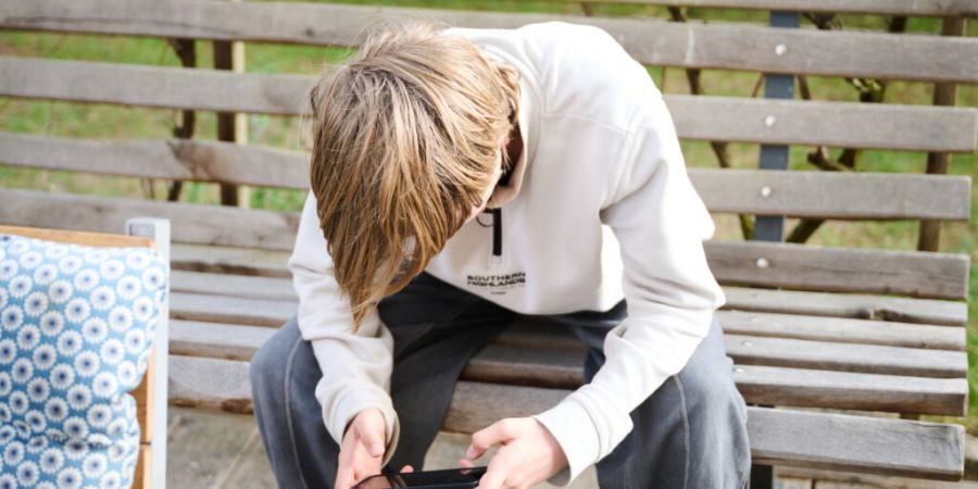 ILLUSTRATION - Ein Jugendlicher sitzt auf einer Bank und schaut auf sein Smartphone. Australien hat ein Social-Media-Verbot für Kinder und Jugendliche unter 16 Jahren eingeführt und i...