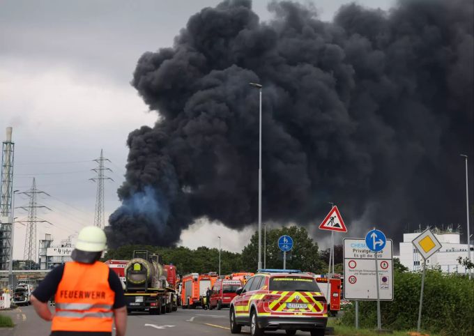 27.07.2021, Nordrhein-Westfalen, Leverkusen: Einsatzfahrzeuge der Feuerwehr stehen unweit einer Zufahrt zum Chempark über dem die dunkle Rauchwolke aufsteigt. Rauchwolke über Chempark Leverkusen