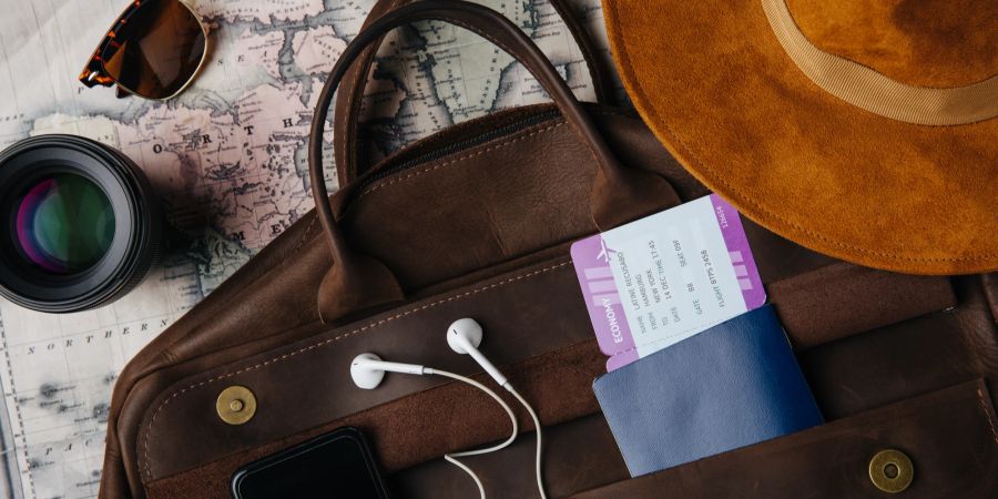Auf einem Tisch liegen eine Landkarte, Hut, Fotolinse und eine Reisetasche mit Ticket, Handy, Kopfhörern.
