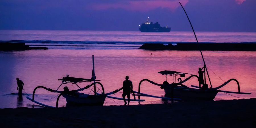 Männer befestigen vor Sonnenaufgang am Tag vor Beginn des G20-Gipfels am Strand von Nusa Dua Boote, wobei im Hintergrund ein Schiff der indonesischen Marine zu sehen ist.