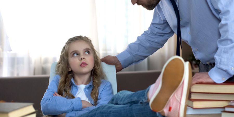 Junges Mädchen an einem Schreibtisch, Arme verschränkt, Beine auf dem Tisch, schaut zum Vater