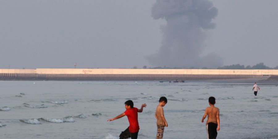 ARCHIV - Drei Jungen spielen im seichten Wasser der Straße von Hormus, während im Hintergrund vor der Küste von Bandar Abbas im Iran eine Rauchwolke nach einer Explosion aufsteigt. F...