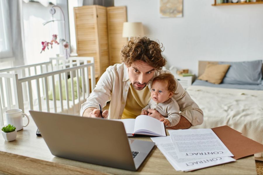 Vater am Laptop mit Baby auf dem Arm