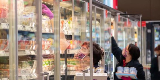 Mitarbeiterinnen in einem Supermarkt tragen Schutzmasken, während sie die Kühlregale mit Waren bestücken. lebensmittel