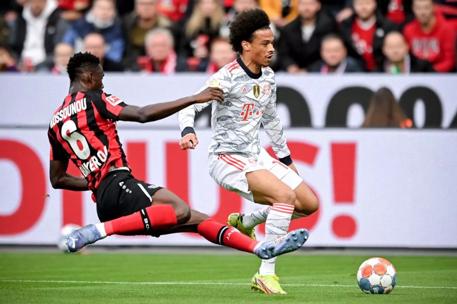 Leverkusens Odilon Kossounou (l.) grätscht gegen Bayern-Flügel Leroy Sané (r.).