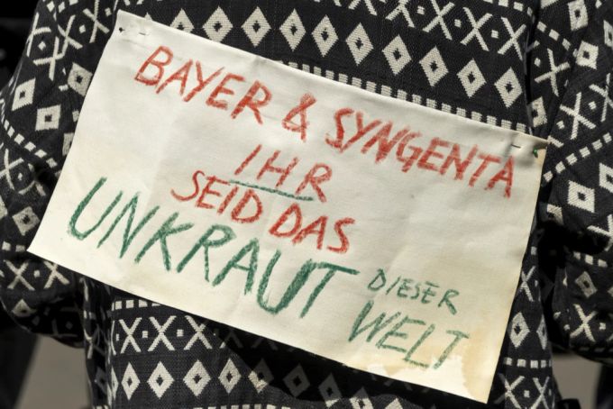 Eine Teilnehmerin der Kundgebung March against Bayer & Syngenta hält ein Plakat mit der Aufschrift BAYER UND SYNGENTA IHR SEID DAS UNKRAUT DIESER WELT vor dem Sitz der Syngenta in Basel Syngenta