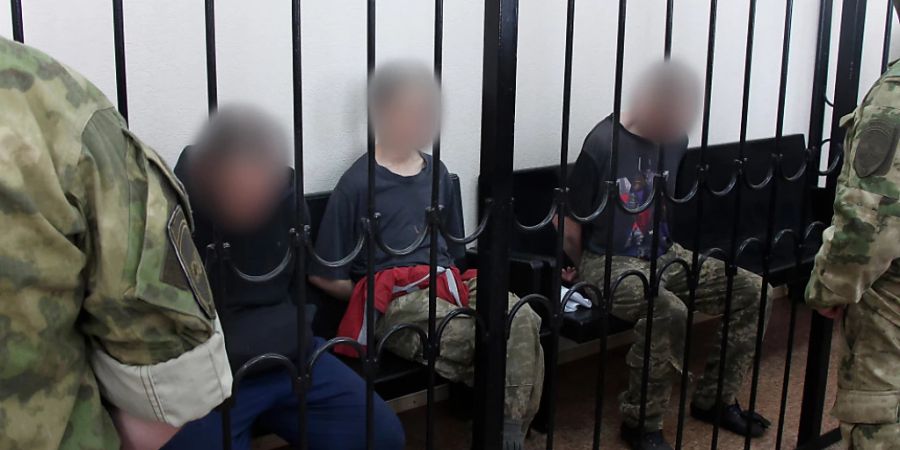 Zwei britische Staatsbürger (links und rechts) sowie ein marokkanischer Staatsbürger sitzen hinter Gittern in einem Gerichtssaal in Donezk. Foto: Uncredited/AP/dpa - ACHTUNG: Personen wurden aus rechtlichen Gründen gepixelt