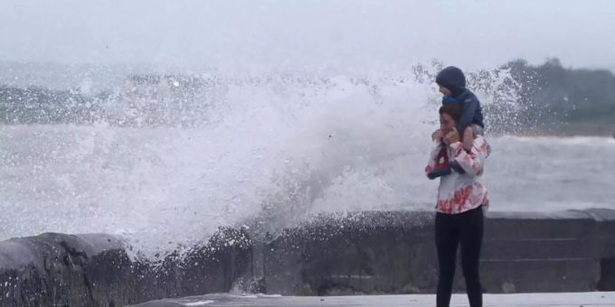 Eine Welle tritt über das Ufer während eine Frau mit einem Kind auf den Schultern an der Strandpromenade geht. Sturmtief "Ellen" trifft auf die Küste Irlands. Foto: Niall Carson/PA Wire/dpa Eine Welle tritt über das Ufer während eine Frau mit einem Kind auf den Schultern an der Strandpromenade geht. Sturmtief "Ellen" trifft auf die Küste Irlands. Foto: Niall Carson/PA Wire/dpa