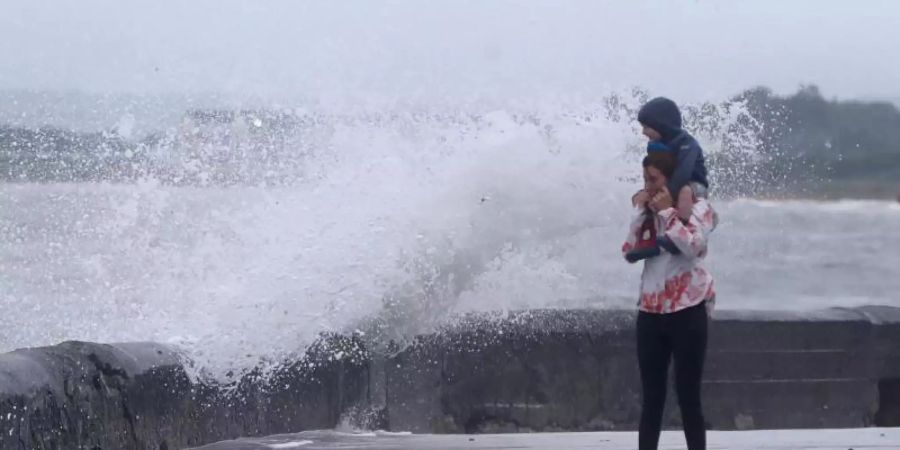 Eine Welle tritt über das Ufer während eine Frau mit einem Kind auf den Schultern an der Strandpromenade geht. Sturmtief "Ellen" trifft auf die Küste Irlands. Foto: Niall Carson/PA Wire/dpa