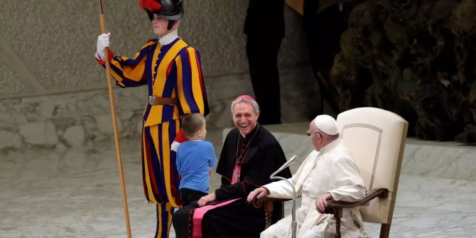 Papst Franziskus und Erzbischof George Gänswein lachen gemeinsam während ein Kind neben ihnen mit einem Mitglied der päpstlichen Schweizergarde spielen möchte.