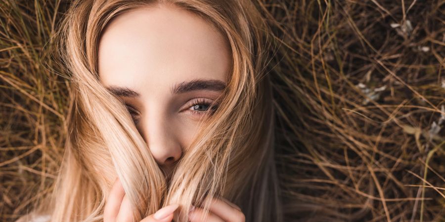 Draufsicht der jungen blonden Frau, die in die Kamera schaut, während sie auf grünem Gras liegt und ihr Gesicht mit Haaren verdeckt.