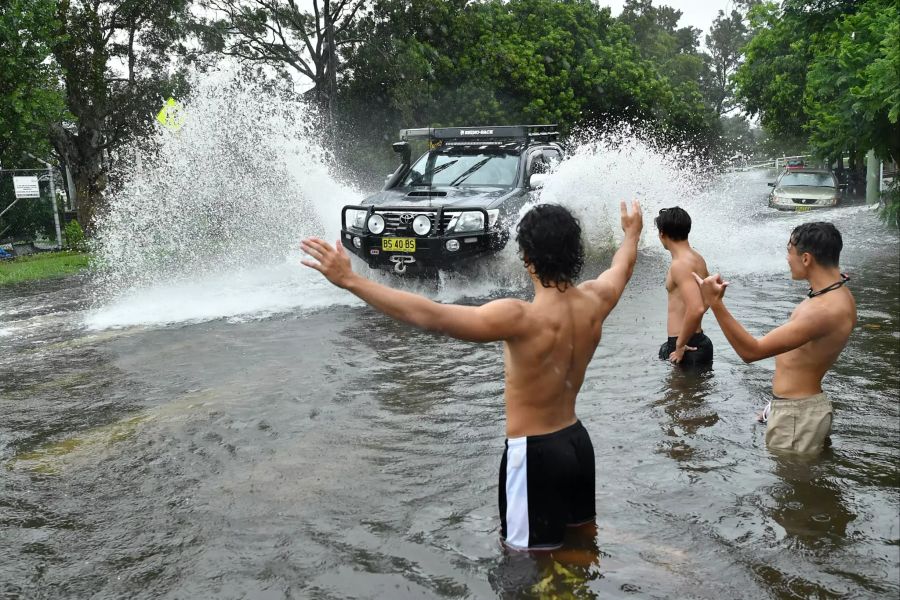 Junge Männer stehen in Sdney auf eine überfluteten Strasse und grüssen ein vorbeifahrendes Auto. Auch in den kommenden Tagen sind noch schwere Unwetter prognostiziert.