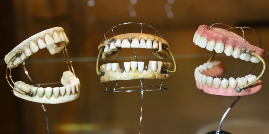 Links ein vollständiges Gebiss aus Elfenbein geschnitzt mit einer Doppelfeder versehen aus der Zeit um 1750, in der Mitte ein sogenanntes Waterloo Gebiss, welches einem Menschen etwa 5 bis 10 Jahre seine Dienste geleistet hat und rechts ein Kautschuckgebiss mit Federn um 1870.