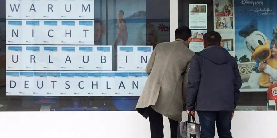 Zwei Männer schauen in das Schaufenster eines Reisebüros, an dem mit grossen Buchstaben «Warum nicht Urlaub in Deutschland» zu lesen ist. Foto: Bernd Thissen/dpa