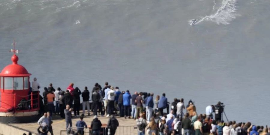 Menschen am Leuchtturm bei Nazaré beobachten einen Surfer am Praia do Norte, der eine Riesenwelle surft. Foto: Armando Franca/AP/dpa