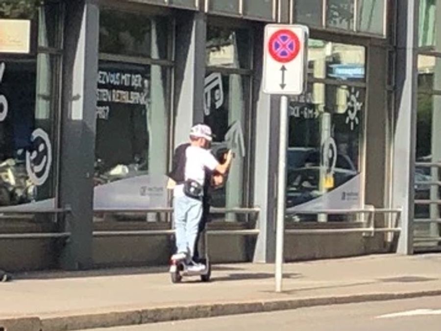 Das ist gemeint: Zwei junge Menschen fahren im Zürcher Kreis 1 in der Nähe des Plakats der Stapo zu zweit auf einem E-Trottinett.