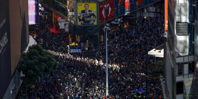 08.12.2019, China, Hongkong: Ein Sonnenstrahlenbündel scheint durch die hohen Häuser auf die pro-demokratischen Demonstranten, die auf einer Strasse durch das Zentrum der Stadt marschieren. Sechs Monate nach Beginn der Proteste gegen die Regierung und für mehr Demokratie hat heute in Hongkong die grösste Demonstration seit Wochen begonnen. Foto: Vincent Thian/AP/dpa +++ dpa-Bildfunk +++ Proteste in Hongkong