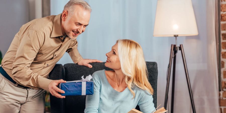 Frau sitzt mit Buch und lächelt neben Ehemann mit Geschenkbox in der Hand.