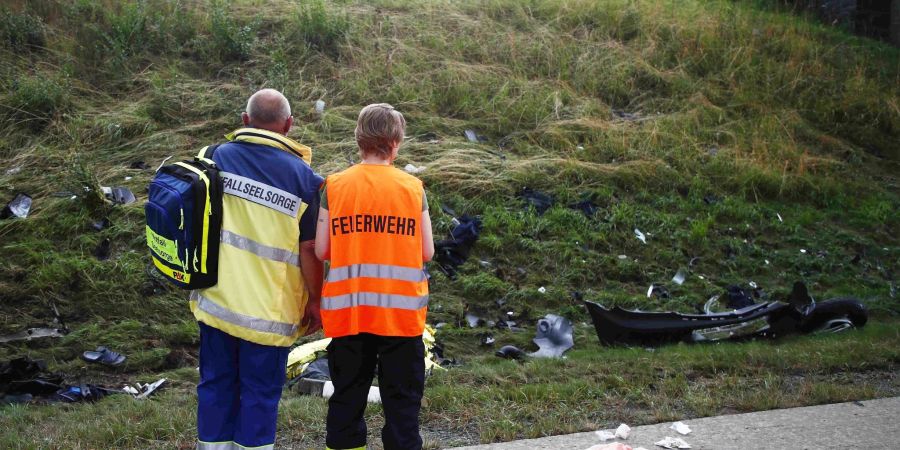 Ein Seelsorger und ein Mitglied der Feuerwehr an einer Unfallstelle auf der Autobahn 9 vor einer Böschung, die mit Trümmerteilen übersäht ist.