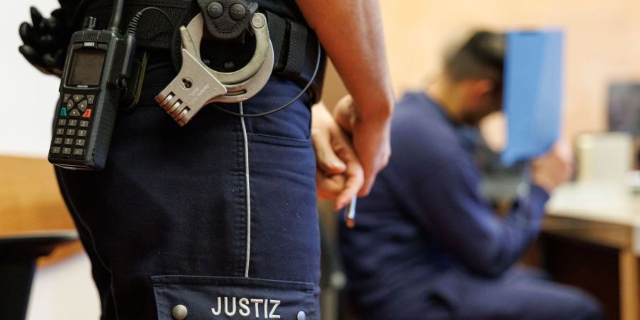 Ein Justizbeamter bewacht den Angeklagten, der sein Gesicht hinter einem Aktendeckel verbirgt.