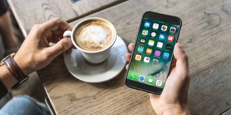 Ein Mann hält ein Smartphone und sitzt dabei an einem Tisch mit einem Kaffee.