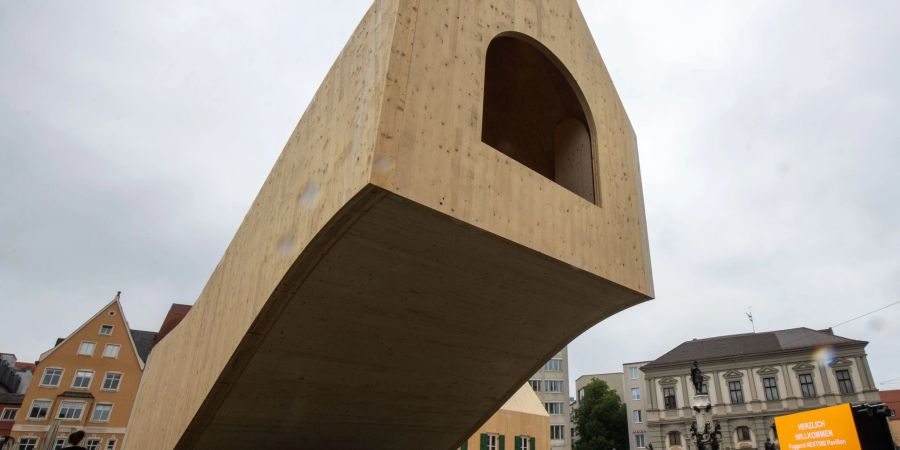 Ein Holzhaus steht freischwebend neben dem Augustusbrunnen. In dem Pavillon auf dem Rathausplatz in Augsburg sollen mehrere Wochen lang zahlreiche Veranstaltung aus Anlass des Jubiläums der ältesten Sozialsiedlung der Welt stattfinden.