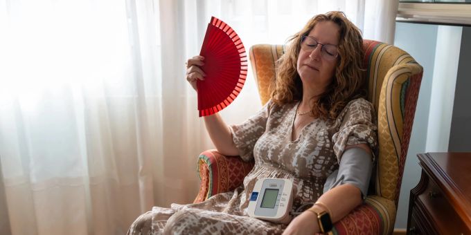Menopause oder Bluthochdruck oder beides? Unterschätzen Sie die Symptome nicht! Frau mit Fächer und Bluthochdruckmessgerät in einem Sessel