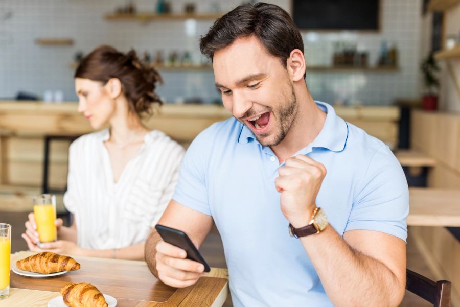 frau schaut betreten zur seite, mann schaut jubilierend auf smartphone