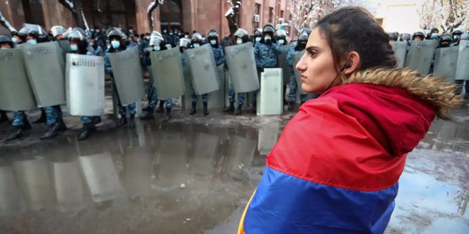 Eine Demonstrantin, die eine armenische Fahne als Umhang trägt, steht vor einer Kette Polizisten mit Schildern während einer Kundgebung fest. Proteste in Armenien