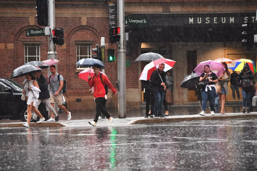 Passanten gehen mit Schirmen durch den Regen. Heftiger Regen hat einige Teile von Australien überflutet, aber auch die Arbeit der Feuerwehr in den Brandgebieten erleichtert.