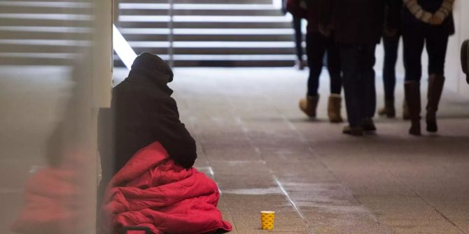 Ein Bettler sitzt in einer Unterführung während Passanten vorbeilaufen.