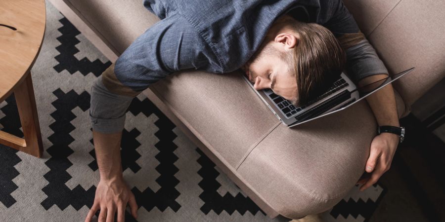 Mann schläft auf der Couch über der Tastatur seines Laptops ein.