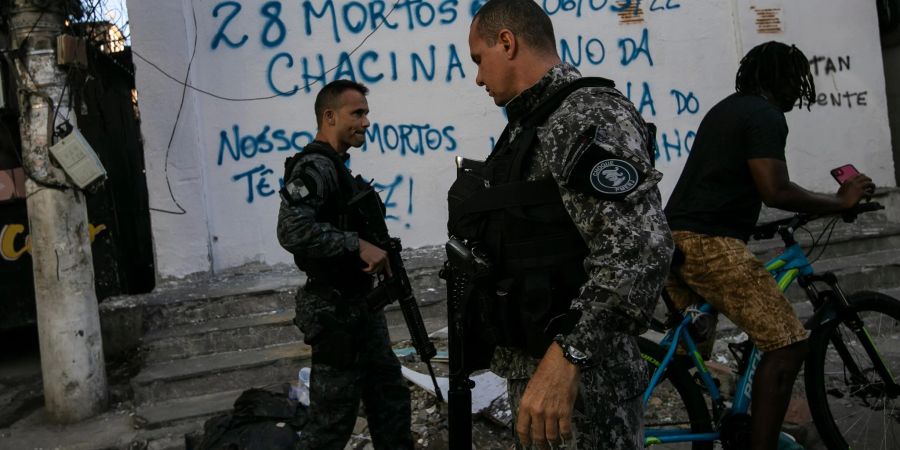 Bereitschaftspolizisten patrouillieren in Rio De Janeiro. Auf der Mauer hinter ihnen steht in portugiesischer Sprache: "28 Tote sind ein Massaker! Unsere Toten haben eine Stimme! Ein Jahr Jacarezinho-Massaker".