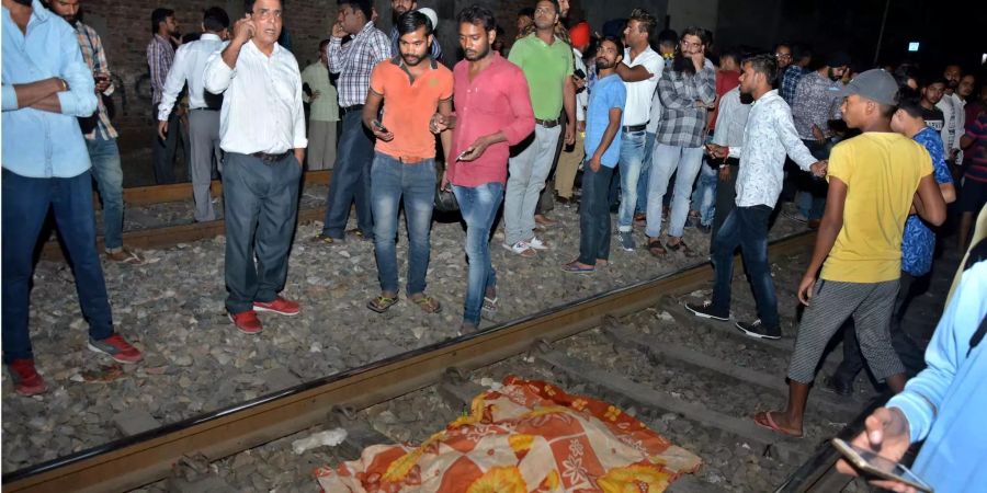 Ein mit einem Tuch abgedeckter Körper liegt nach einem Zugunglück auf Bahnschienen, während indische Männer auf und neben den Schienen stehen.