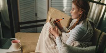 Junge Frau mit Decke auf Sofa malt oder schreibt in ein Heft