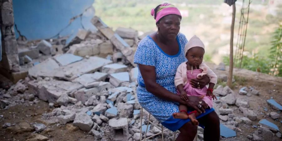 Eine Frau sitzt mit einem Kind vor einem zerstörten Haus in Haiti.