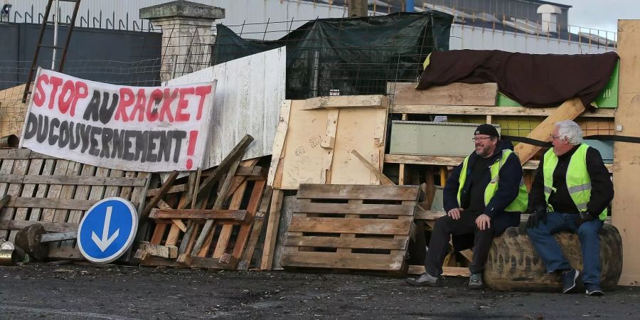 Demonstranten sitzen vor einer provisorischen Barrikade der «Gelbwesten», an der ein Banner mit der Aufschrift «Stop au racket du gouvernement!» (Stoppt die Erpressung der Regierung) hängt.