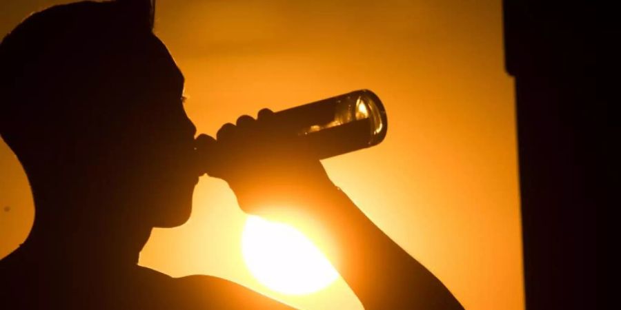 Ein junger Mann sitzt auf dem Kronsberg, und trinkt Bier, während am Horizont die Sonne untergeht. Foto: Julian Stratenschulte/dpa