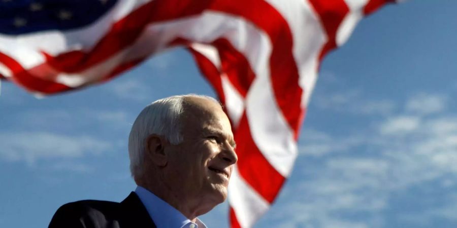 John McCain lächelt im Vordergrund einer amerikanischen Flagge, die im Wind weht.