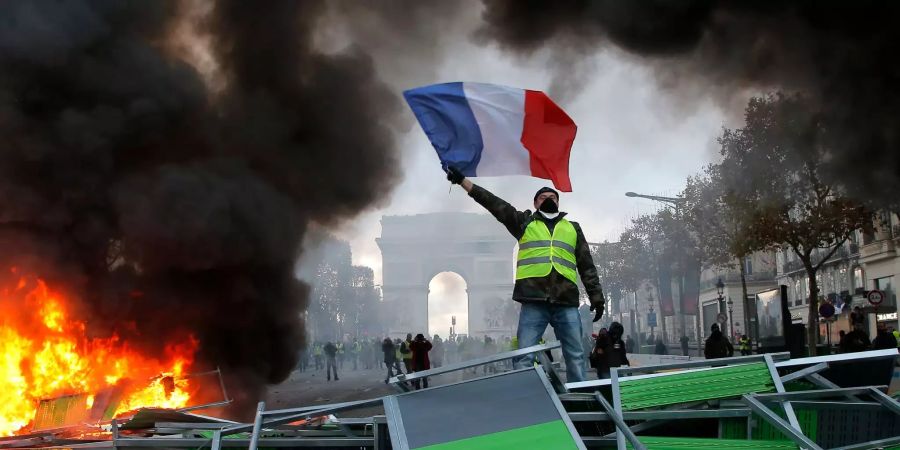 Ein Demonstrant mit einer gelben Weste schwenkt die französische Flagge neben einer brennenden Barrikade auf der Prachtstrasse Champs Elysee mit dem Arc de Triomphe im Hintergrund.