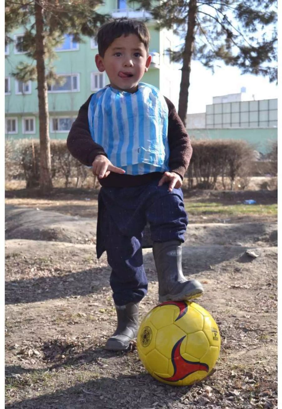 Der kleine Murtasa Ahmadi steht in Gummistiefeln und in einem Lionel-Messi-Trikot aus einer Plastiktüte auf einem Platz.
