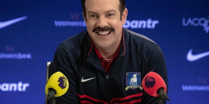 Jason Sudeikis als Ted Lasso in der gleichnamigen Serie von Apple TV+. Jason Sudeikis vor Mikrofonen.