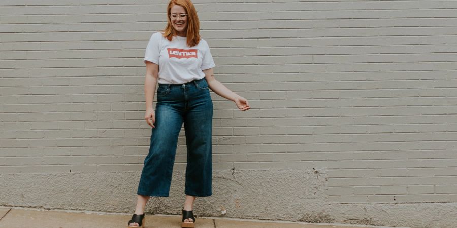 Frau mit roten Haaren, Brille, Lentils Shirt und Jeans auf der Strasse vor einer grauen Mauer.