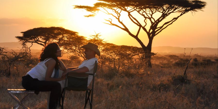 Paar sitzt in der Savanne vor untergehender Sonne umgeben von Bäumen und afrikanischer Wildnis.