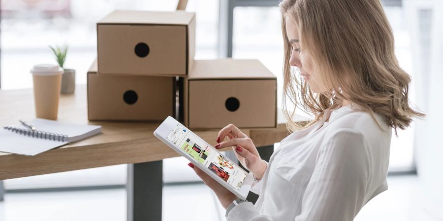 Eine Frau sitzt vor einem mit Päckchen bestückten Schreibtisch und tippt auf ein Tablet.