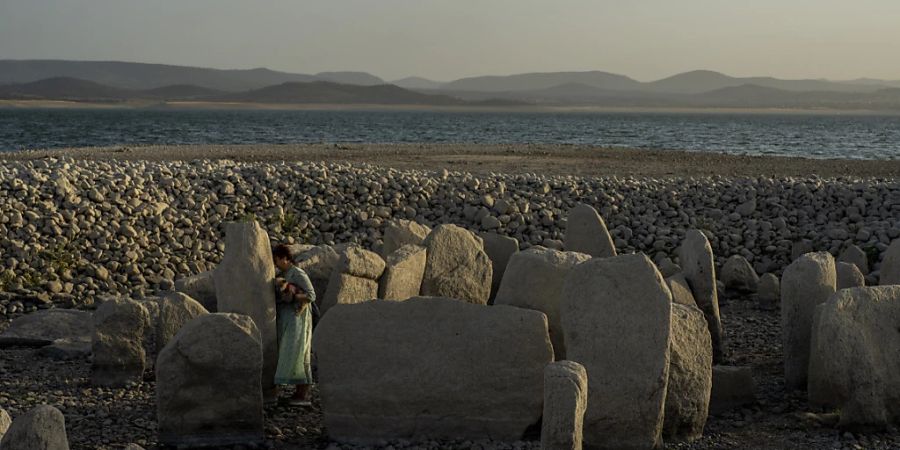 Die 54-jährige Amalie Garcia geht mit einem Hund im Arm an den Dolmen von Guadalperal vorbei, einem megalithischen Monument, das aufgrund der Dürre am Valdecanas-Stausee zu sehen ist. Foto: Manu Fernandez/AP/dpa