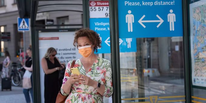 Eine Schwedin mit Schutzmaske vor einem Schild, dass aufs Social Distancing aufmerksam macht. Coronavirus in Stockholm