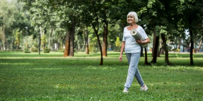 Wie Sie auch im Alter fit bleiben, erfahren Sie hier. Ältere Frau im Park mit Yogamatte.